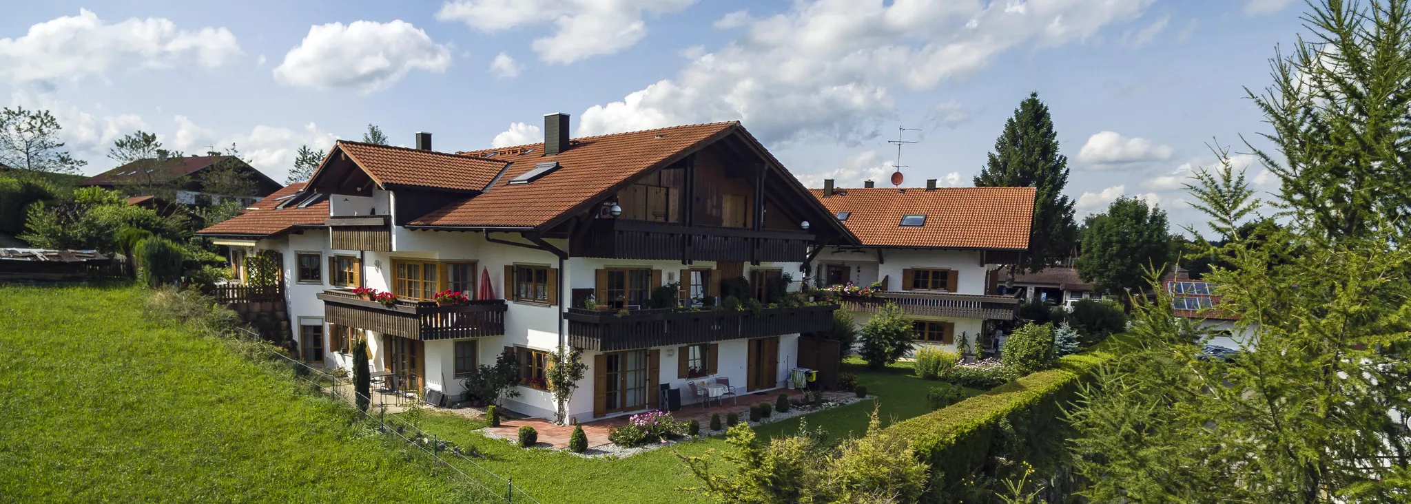 Ein großes, zweistöckiges Haus mit einem schrägen roten Dach und Holzbalkonen ist von einem üppigen Rasen und Büschen umgeben. Der Himmel ist teilweise bewölkt und im Hintergrund sind die Nachbarhäuser zu sehen.