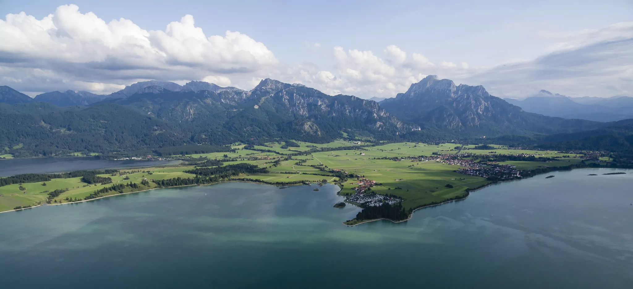 Luftaufnahme des Forggensees, umgeben von grünen Feldern und kleinen Dörfern. Im Hintergrund erheben sich majestätische Berge unter einem teilweise bewölkten Himmel.