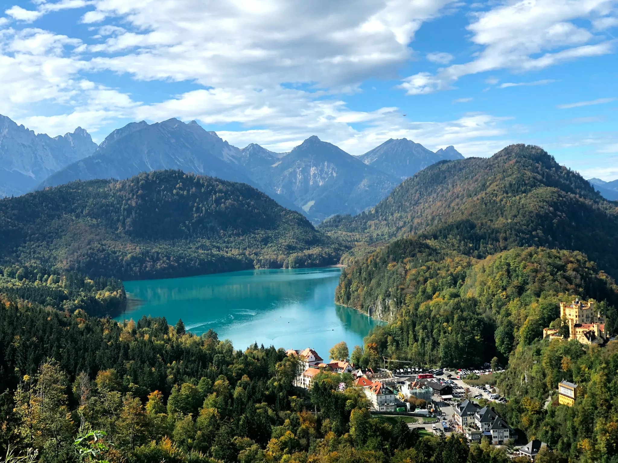Eine malerische Aussicht auf einen türkisfarbenen See, umgeben von üppigen, bewaldeten Bergen unter einem blauen Himmel mit vereinzelten Wolken. Am Fuße der Hügel ist ein malerisches Dorf mit einer Burg zu sehen. Im Hintergrund erheben sich majestätische Gipfel.