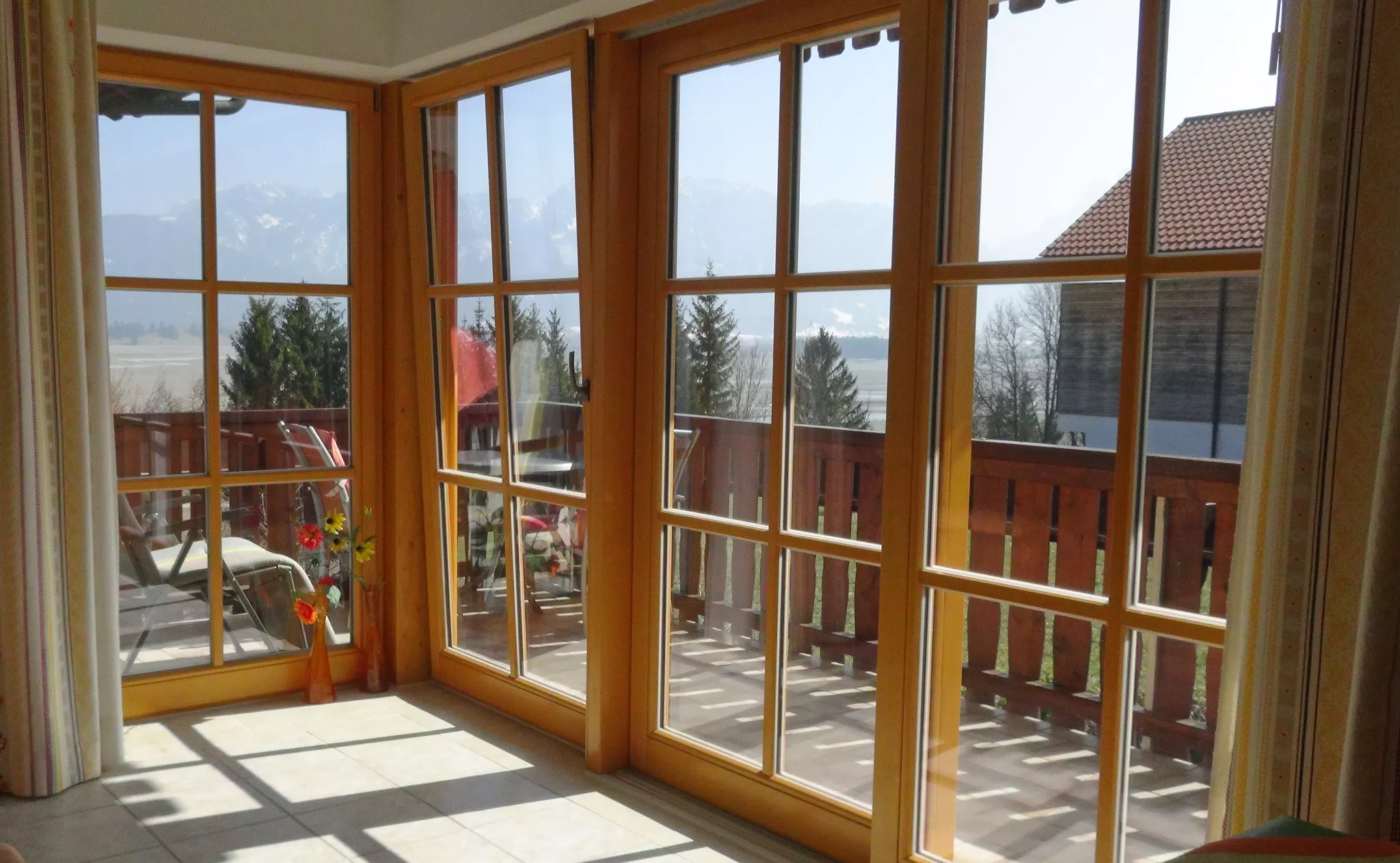Ein sonnendurchfluteter Raum mit großen Glastüren in Holzrahmen öffnet sich zu einem Balkon mit Blick auf die Berge in der Ferne. Das Sonnenlicht wirft Muster auf den gefliesten Boden und in der Ecke steht eine Vase mit Blumen, die einen Hauch von Farbe verleiht.