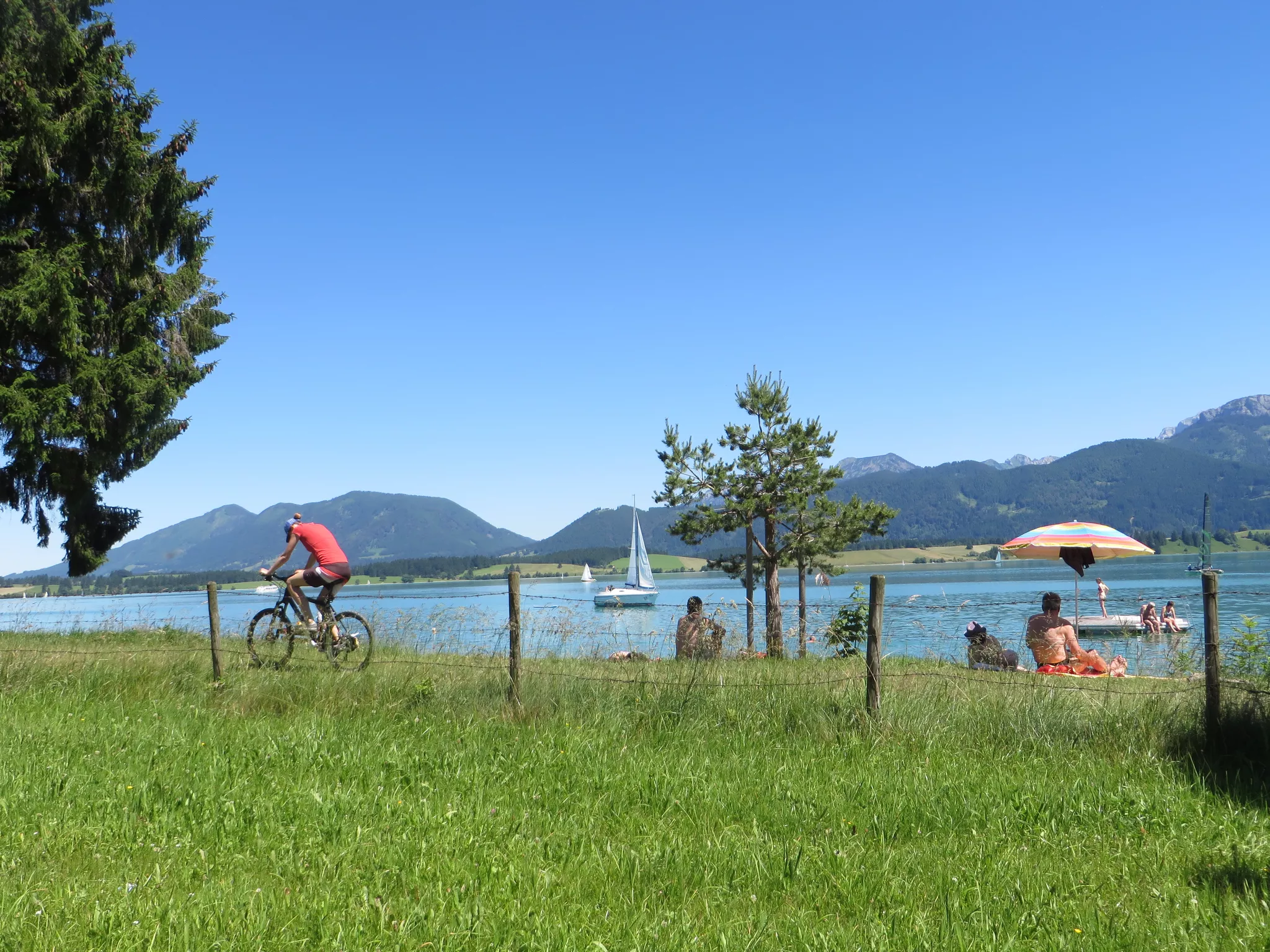 Ein Radfahrer fährt auf einem Grasweg in der Nähe eines Sees mit Segelbooten. Menschen sitzen am Wasser, einige unter einem Sonnenschirm. Um ihn herum sind Berge und Bäume unter einem klaren blauen Himmel.